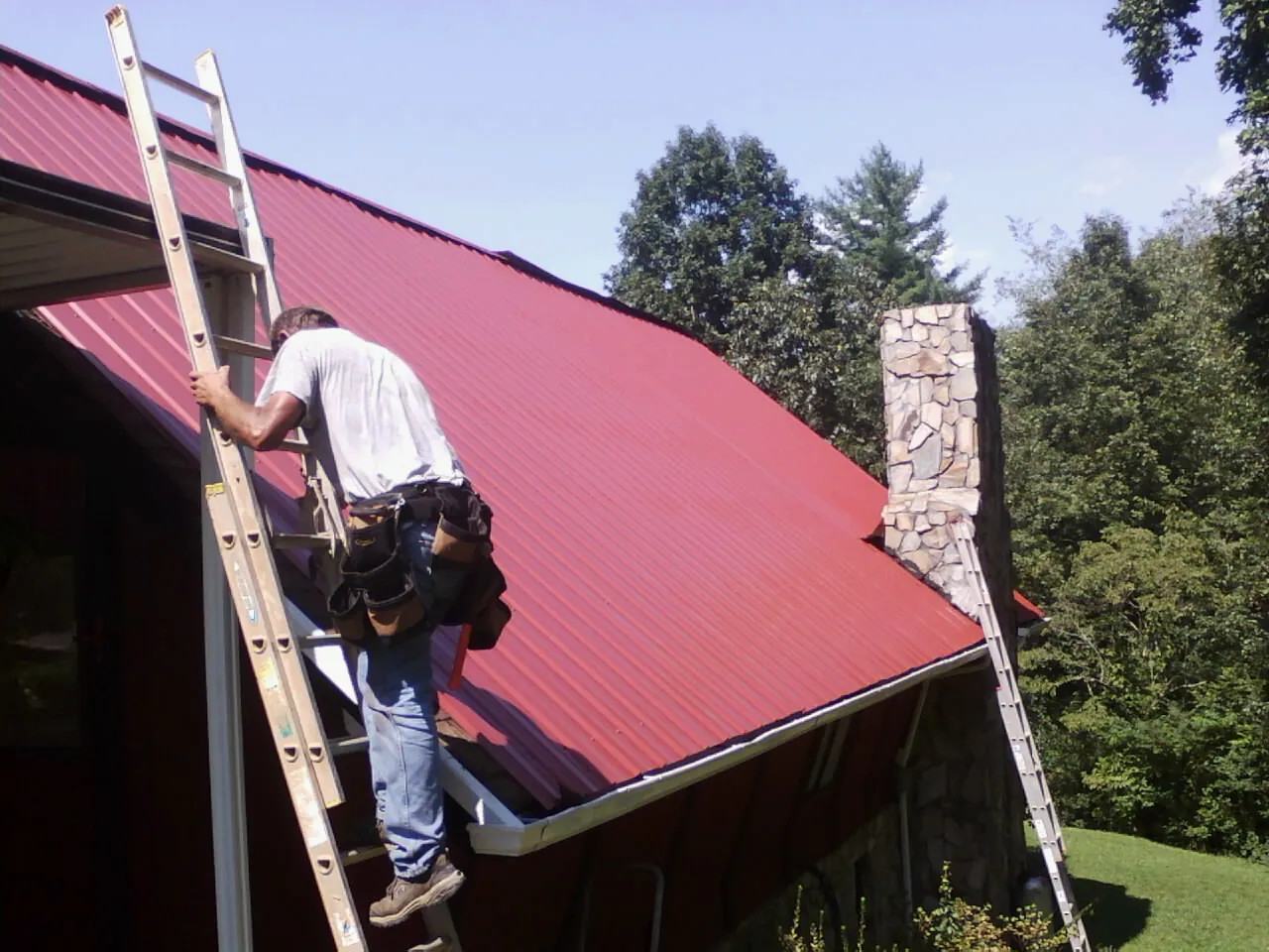 Roofer on a ladder beside a red metal roof and stone chimney — Soffit & Fascia Repair by Roofing Brothers in Hemingway